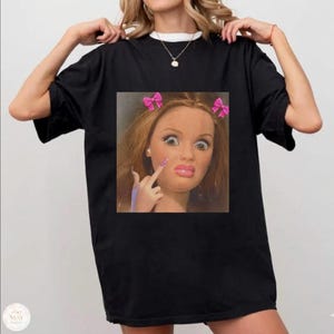 Puede incluir: Camiseta negra con un gráfico cuadrado de una cara de muñeca con lazos rosas, uñas largas y un gesto con el dedo. La camiseta tiene un cuello blanco y la lleva una persona.