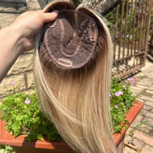 Może przedstawiać: Blond i jasnobrązowy topper do włosów z brązową podstawą i kilkoma klipsami do mocowania. Peruka jest trzymana w górze, prezentując naturalnie wyglądającą mieszankę kolorów i konstrukcję czepka.