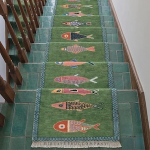 Könnte beinhalten: Ein grüner Treppenläufer mit einem sich wiederholenden Muster aus stilisierten Fischen in verschiedenen Farben wie Orange, Blau, Rosa und Gelb. Der Teppich hat eine blaue geometrische Bordüre und weiße Fransen mit dem Text MIRZAPURRUGCOMPANY.
