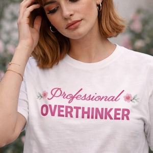May include: White t-shirt with the text "Professional Overthinker" in pink script. The text is decorated with pink flowers and green leaves. The shirt is a crew neck style.
