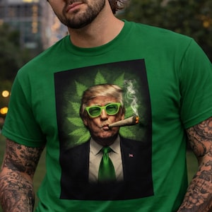 Könnte beinhalten: Grünes T-Shirt mit einer Grafik eines Mannes, der eine grüne Sonnenbrille trägt und eine brennende Zigarre raucht. Die Grafik enthält ein grünes Cannabisblatt-Design im Hintergrund. Der Mann trägt eine grüne Krawatte und einen schwarzen Anzug.