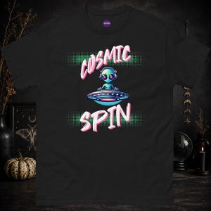 Könnte beinhalten: Ein schwarzes T-Shirt mit einer Grafik eines blauen Außerirdischen, der Kopfhörer trägt und eine fliegende Untertasse als Plattenspieler bedient. Der Text "COSMIC SPIN" ist in Pink über und unter der Illustration auf grünem Punktmuster.