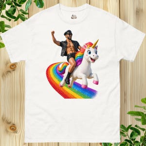 Puede incluir: Una camiseta blanca con un estampado gráfico de una persona con chaleco, pantalones cortos y gorra de cuero negro, montando un unicornio blanco con crin y cola de arcoíris sobre un camino multicolor.