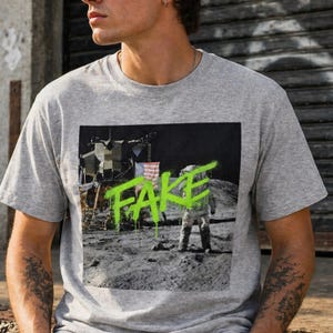 Puede incluir: Camiseta gris de cuello redondo con un gráfico del alunizaje, incluyendo un astronauta y la bandera estadounidense. La palabra "FAKE" está escrita en pintura en aerosol verde neón sobre la imagen.
