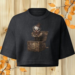 Könnte beinhalten: Ein schwarzes bauchfreies T-Shirt mit einem Grafikdruck einer verwitterten, braunen Spieluhr, aus der ein unheimlicher Clown hervorspringt. Der Clown trägt einen spitzen Hut und einen gerüschten Kragen. An der Box befindet sich eine Metallkurbel.