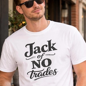 Puede incluir: Camiseta blanca con la frase "Jack of NO Trades" en letras negras. La camiseta es de cuello redondo y parece ser de algodón. El texto está centrado en la parte delantera de la camiseta.