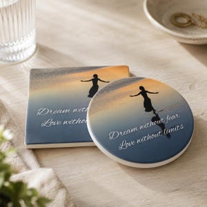 May include: A square and a round ceramic coaster featuring a sunset beach scene with a silhouette of a person dancing. The text reads Dream without fear, Love without limits. The coasters have a gradient from dark blue to orange and yellow.