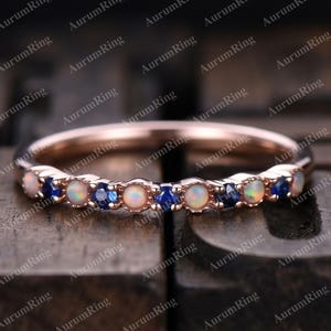 May include: A rose gold band ring featuring an alternating pattern of round white opals and small round blue sapphires set along the front half of the band. The metal has a polished finish, and the stones are arranged in a repeating sequence.