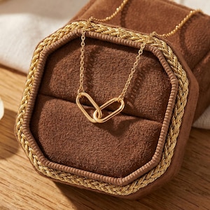 Puede incluir: Un collar de tono dorado con dos eslabones ovalados entrelazados como colgante central, que descansa sobre un joyero forrado de terciopelo marrón con un borde trenzado dorado. La delicada cadena se extiende desde el colgante por la caja.