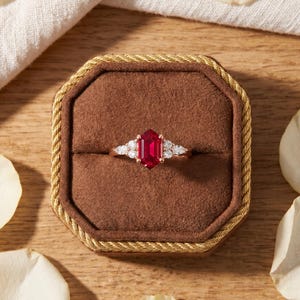 May include: A gold ring featuring a central hexagonal red gemstone flanked by three small clear stones on each side. The ring is displayed in a brown octagonal velvet box with a gold braided trim, surrounded by scattered white flower petals.