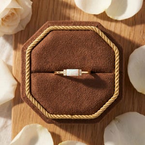 May include: A gold ring featuring a rectangular opal stone set horizontally. The opal is flanked by two small, clear accent stones on each side. The ring is displayed centered within a brown, octagonal, velvet-lined jewelry box with gold trim.