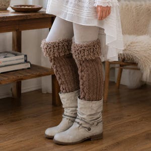 May include: A pair of brown knit leg warmers featuring a vertical cable knit pattern and a textured, looped cuff at the top. The leg warmers are worn over light-colored leggings and paired with light gray slouchy boots that have a decorative chain detail.