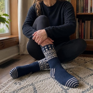 May include: A pair of mid-calf knit socks featuring a deep navy blue base with a contrasting cream-colored geometric pattern around the cuff and ankle. The toe and heel sections are detailed with a fine navy and cream striped ribbed texture.