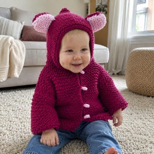 May include: A deep magenta knitted hooded jacket featuring light pink rounded ears on top of the hood and four light pink toggle closures down the front. The garment is crafted with a thick, textured knit stitch and long sleeves for warmth.