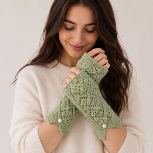 May include: A pair of sage green fingerless gloves featuring an intricate knit lace pattern with small bobbles. Each glove has a scalloped edge at the wrist adorned with a single white pearl accent, providing a textured and detailed appearance.
