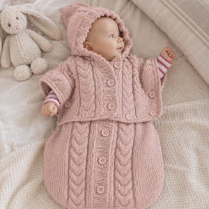 May include: A dusty pink knitted baby sleeping bag with a matching hooded capelet. The set features a vertical cable knit pattern, a front button closure on both the capelet and the sleeping bag, and short sleeves. A plush cream-colored rabbit toy is nearby.