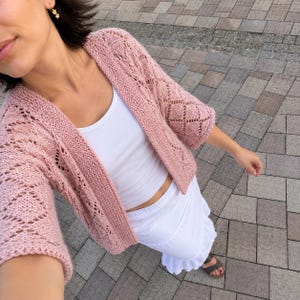 May include: A dusty pink open-front cardigan featuring a delicate diamond-shaped knit pattern with three-quarter length sleeves, worn over a plain white tank top and a white tiered skirt with a ruffled hemline and dark grey open-toe sandals.