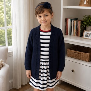 May include: A navy blue open-front knit cardigan with a textured border pattern, worn over a white and navy blue horizontal striped dress featuring a red neckline trim. A dark blue fabric flower headband is worn on the head.