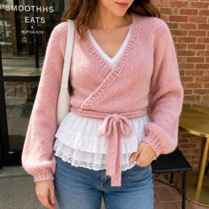 Könnte beinhalten: Ein weicher, rosa Strick-Wickelpullover mit langen Ärmeln und Bindegürtel, getragen über einem weißen Unterhemd mit gestuftem Rüschensaum und Lochstickerei. Der Pullover hat einen V-Ausschnitt und wird mit blauen Jeans und einer weißen Tasche kombiniert.