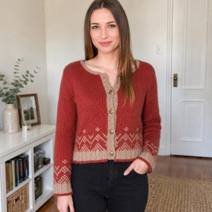 May include: A rust-coloured knit cardigan featuring a round neckline, long sleeves, and a front button closure. The garment is accented with a tan-coloured trim along the placket, cuffs, and hem, which includes a repeating geometric zigzag pattern.