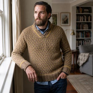 Könnte beinhalten: Ein brauner Strickpullover mit einem strukturierten Muster mit kleinen gelben Akzenten, einem Schalkragen und gerippten Bündchen. Der Pullover wird über einem hellblauen Hemd mit Kragen und einer dunkelblauen Jeans getragen und hat lange Ärmel.