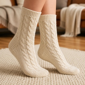 May include: A pair of cream-colored knit socks featuring a vertical cable knit pattern running along the front. The socks have a ribbed cuff at the top and a reinforced heel and toe section, providing a textured appearance and a snug fit.