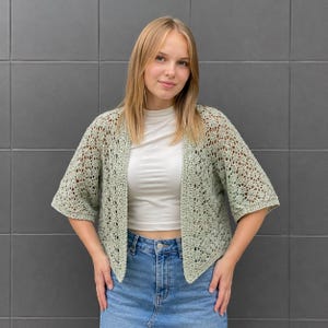 May include: A sage green open-front crochet cardigan featuring a delicate, intricate lace-like pattern throughout the body and sleeves. The garment has a relaxed fit with three-quarter length sleeves and a straight hemline, worn over a white top.