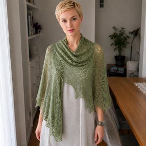 Könnte beinhalten: Ein olivgrünes, dreieckiges Tuch mit einem komplizierten, durchbrochenen Spitzenmuster und einem zarten, gewellten Rand. Das leichte Strickaccessoire liegt über einem schlichten, hellen ärmellosen Kleidungsstück und zeigt seine detaillierte Struktur.
