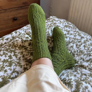 Op de afbeelding: Een paar sokken tot halverwege de kuit in een levendige olijfgroene kleur. De sokken hebben een verticaal kabelbreipatroon aan de voor- en zijkanten, met een geribbelde boord aan de bovenkant en een verstevigde hiel en teen in hetzelfde garen.