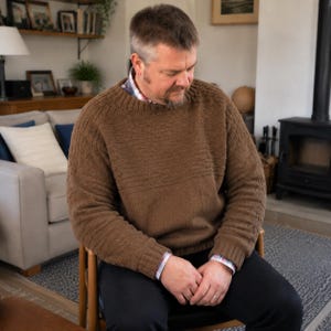May include: A brown long-sleeved knit sweater featuring a ribbed crew neckline, cuffs, and hem. The garment displays a textured pattern across the chest and sleeves, layered over a collared shirt with a visible plaid pattern at the neck and wrists.