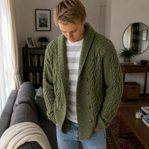 May include: An olive green cable knit cardigan featuring a wide shawl collar, long sleeves, and a double-breasted front closure with four round buttons. The garment is worn over a white and grey horizontal striped t-shirt and light blue denim jeans.
