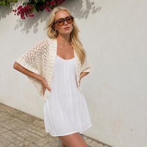Könnte beinhalten: Eine cremefarbene, offene Häkelstrickjacke mit strukturiertem Spitzenmuster und gerippten Kanten, getragen über einem schlichten, ärmellosen weißen Minikleid. Die Strickjacke hat kurze, weite Ärmel und eine leichte, luftige Strickstruktur.