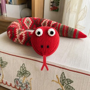 May include: A knitted snake toy featuring a bright red head with white and black button eyes, a red forked tongue, and a body patterned with red, olive green, and cream geometric stripes. The plush creature is coiled and has a textured knit finish.
