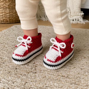 May include: A pair of red and white crocheted baby booties designed to resemble high-top trainers. The booties feature white laces, a white toe cap, a black stripe along the sole, and a small white circular button detail on the outer side.