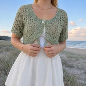 May include: A sage green short-sleeved knit cardigan featuring an open-front design with a single white button closure at the neckline, a ribbed collar, and a delicate lacy openwork pattern throughout the bodice and sleeves.