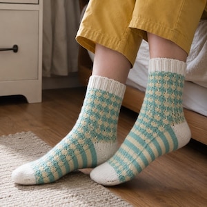 Könnte beinhalten: Ein Paar wadenhohe Strickstrümpfe mit einem cremefarbenen Rippbündchen, einem helltürkis-cremefarbenen Karomuster am Schaft sowie einem helltürkis-cremefarbenen Streifenmuster am Fußteil und einer einfarbigen, cremefarbenen Fußspitze.