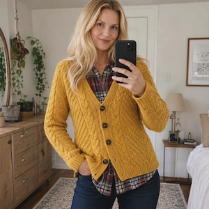 May include: A mustard yellow V-neck cardigan featuring a cable knit pattern on the front panels and sleeves. The garment has a button-down front with four dark brown buttons and ribbed cuffs and hem, worn over a plaid button-up shirt and dark jeans.