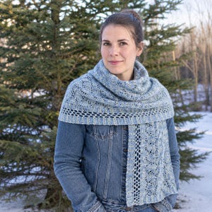 May include: A light blue and white variegated knit scarf featuring an intricate lace pattern with a textured border, draped over a blue denim jacket. The scarf displays a complex openwork design with repeating geometric motifs and ribbed edges.