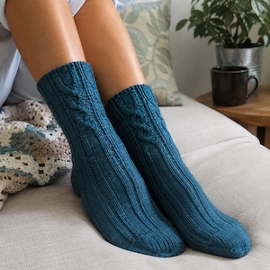 May include: A pair of teal blue knitted socks featuring a vertical cable knit pattern running down the center and ribbed texture along the sides. The socks are crafted from a soft yarn with a subtle shimmer, extending to the mid-calf area.