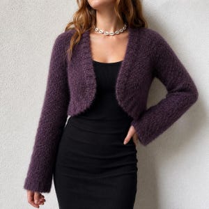 May include: A deep purple, long-sleeved, open-front cropped cardigan with a fuzzy, textured knit finish. The garment features a ribbed trim along the front edges and is worn over a simple black sleeveless dress, paired with a white shell bead necklace.