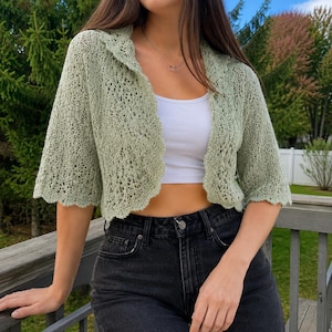 Könnte beinhalten: Eine salbeigrüne, vorne offene Häkelstrickjacke mit kurzen Ärmeln und gewelltem Saum, getragen über einem schlichten weißen bauchfreien Top und einer schwarzen Jeans. Das Strickmuster weist eine filigrane, luftige Textur auf.