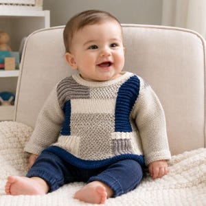 Puede incluir: Un suéter de bebé de manga larga con un diseño de patchwork de bloques de color en crema, azul marino y gris. La prenda de punto tiene un cuello redondo y un patrón texturizado, combinado con pantalones de punto azul marino, usado por un bebé sonriente.