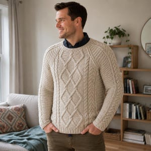 May include: A beige long-sleeve sweater featuring a prominent cable knit pattern on the front and sleeves, a round neckline, and ribbed cuffs and hem. The sweater is worn over a dark collared shirt, paired with olive green trousers.