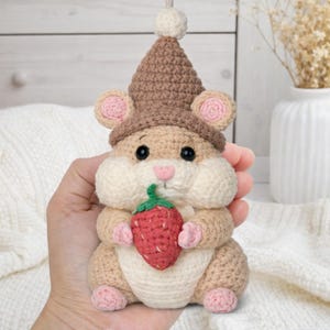 May include: A crocheted hamster doll in beige, cream, and brown, wearing a brown hat and holding a red strawberry. The doll has pink ears, nose, and feet. The hamster is approximately 5 inches tall.