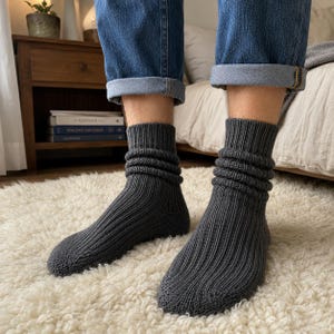 Puede incluir: Un par de calcetines de punto en color gris carbón oscuro con textura acanalada y diseño holgado. Los calcetines llegan por encima del tobillo y se llevan con vaqueros azules con dobladillo, resaltando el patrón de punto vertical del tejido.