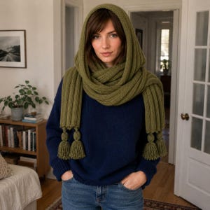 May include: An olive green knitted hooded scarf featuring a thick ribbed texture and long fringe tassels at the ends. The scarf is draped around the head and neck, layered over a dark navy blue long-sleeved sweater and blue denim jeans.