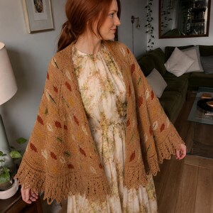 May include: A tan knitted shawl featuring a repeating pattern of small, multi-colored leaf motifs in shades of rust, green, and cream. The shawl has a wide, intricate lace border along the bottom edge and is draped over a light-colored floral dress.