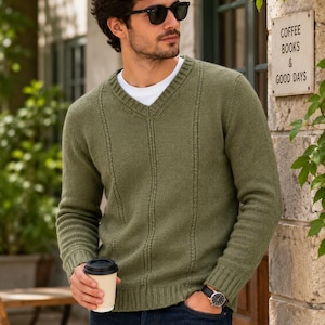 Pode incluir: Um suéter verde-oliva com decote em V, apresentando detalhes canelados verticais na frente, mangas compridas com punhos canelados e uma bainha canelada. O suéter é usado sobre uma camiseta branca lisa de gola redonda e jeans azul-escuro.