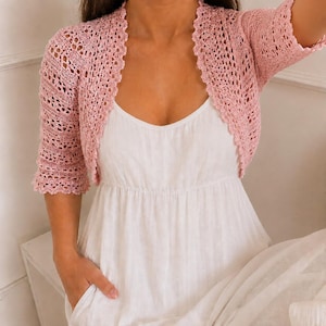 May include: A light pink, open-front crochet bolero with short sleeves and a scalloped edge, worn over a simple white dress. The bolero features an intricate, airy, open-work knit pattern throughout the fabric, providing a delicate texture.