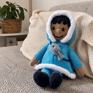 May include: A hand-knitted doll featuring a brown complexion, black hair, and a bright blue hooded coat with white fuzzy trim. The doll wears a small grey fish accessory attached to a black cord around its neck and has black shoes on its feet.